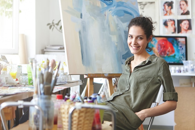 Professional female painter sitting at chair in art studio, keeping hands in pockets of her shirt, smiling gently while resting after drawing picture with watercolors. People, hobby, painting concept