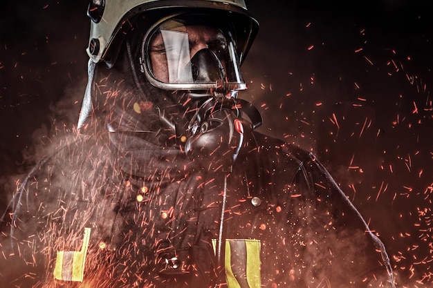 Free photo a professional firefighter dressed in uniform and an oxygen mask standing in fire sparks and smoke over a dark background.