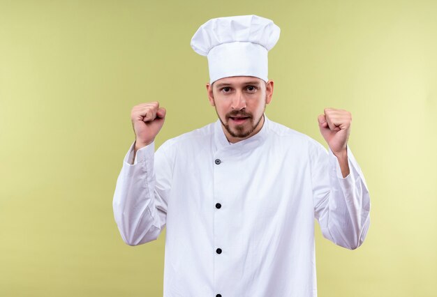 Professional male chef cook in white uniform and cook hat clenching fists happy and excited rejoicing his success standing over green background