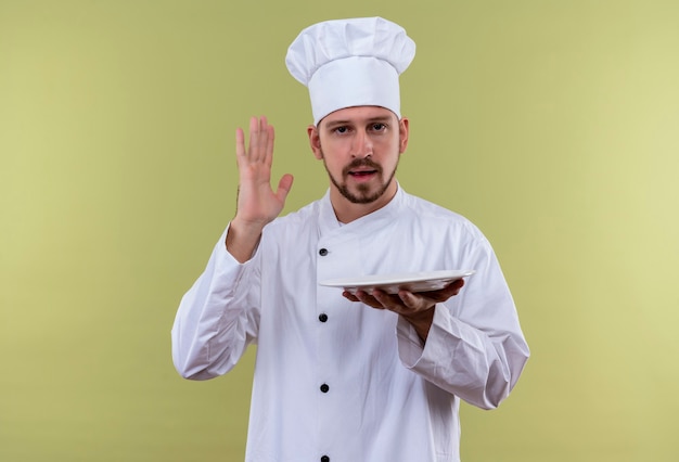 Professional male chef cook in white uniform and cook hat holding an empty plate raising hand standing over green background