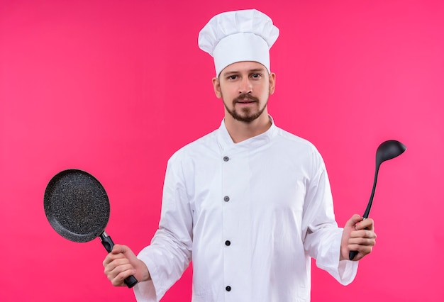 Free photo professional male chef cook in white uniform and cook hat holding a pan and ladle looking at camera with confident smile standing over pink background