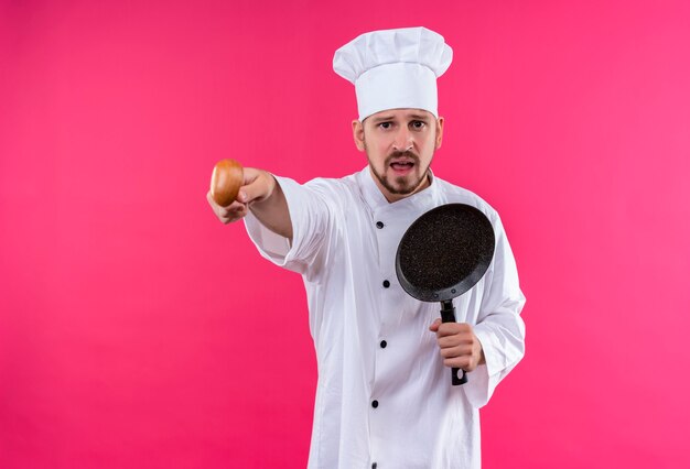 Professional male chef cook in white uniform and cook hat holding a pan pointing to something with a wooden spoon emotional and worried standing over pink background