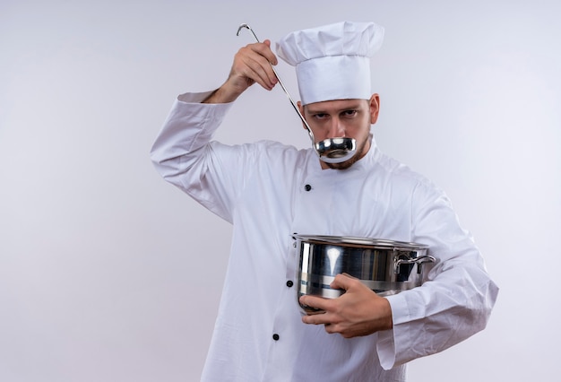 Free photo professional male chef cook in white uniform and cook hat holding a pan tasting food with a ladle standing over white background