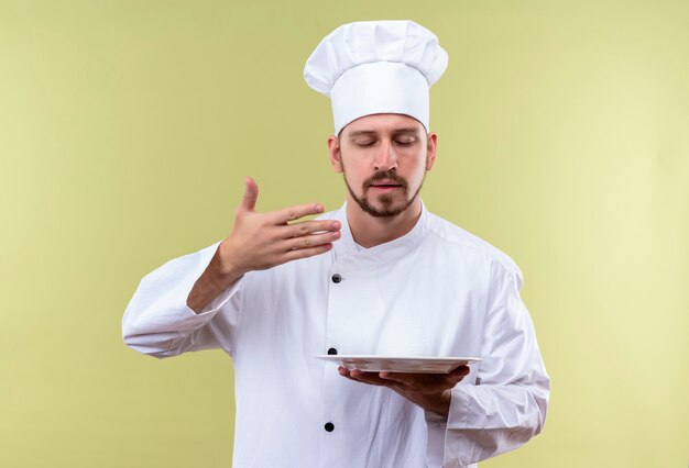 Professional male chef cook in white uniform and cook hat holding plate inhales pleasant smell of food standing over green background