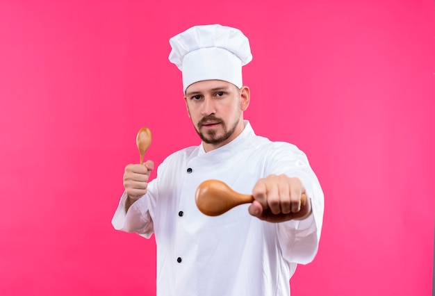 Professional male chef cook in white uniform and cook hat holding wooden spoons looking at camera with serious face standing over pink background