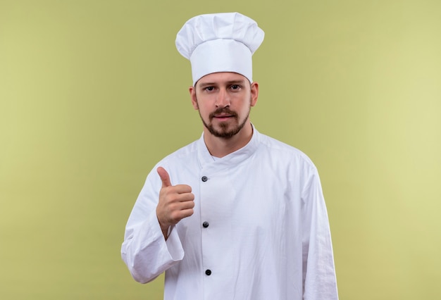 Professional male chef cook in white uniform and cook hat looking at camera showing thumbs up standing over green background