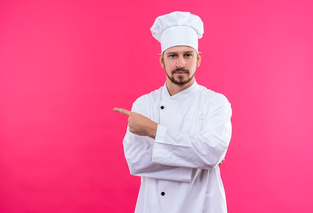 Professional male chef cook in white uniform and cook hat pointing withfinger to the side looking at camera with confident expression standing over pink background