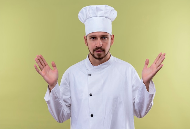 Professional male chef cook in white uniform and cook hat raising hands looking confident standing over green background