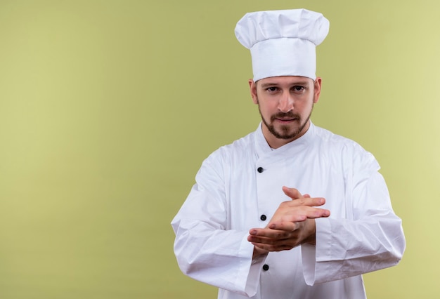 Professional male chef cook in white uniform and cook hat rubbing his hands looking confident, ready to cook standing over green background