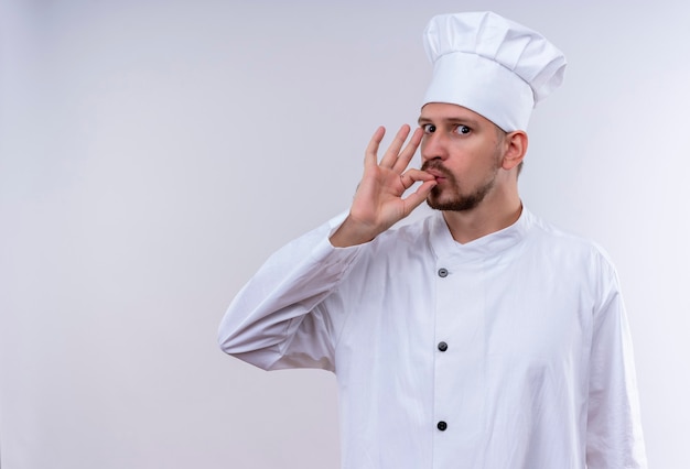 Professional male chef cook in white uniform and cook hat showing sign for delicious standing over white background