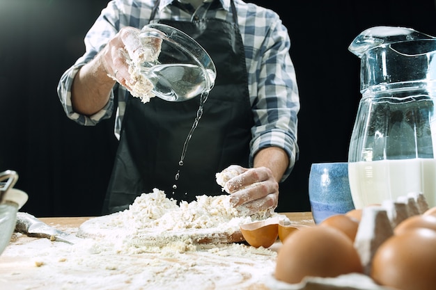 Free photo professional male cook sprinkles dough with flour, preapares or bakes bread or pasta at kitchen table, has dirty uniform, isolated over black chalk background. baking concept