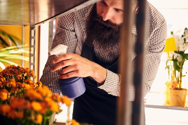 Professional male florist with beard and tattoo on his hand wearing uniform taking care of flowers in a modern flower shop.