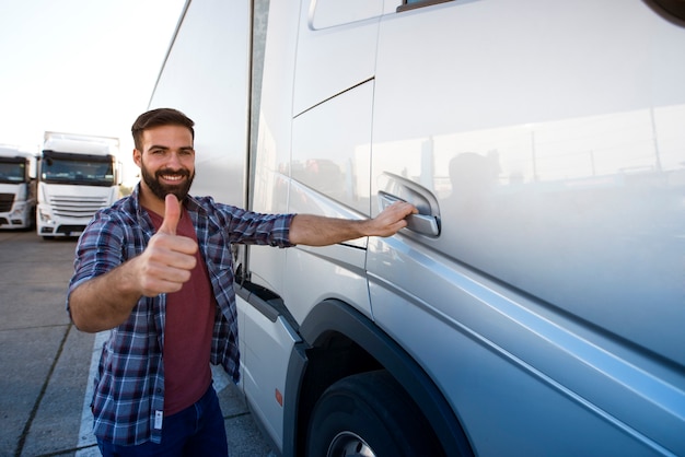 Professional middle aged bearded trucker standing by his semi truck and holding thumbs up