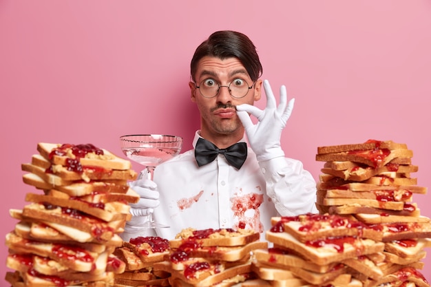 Free photo professional waiter stands with glass of alcoholic cocktail, shows perfect taste sign, has white shirt dirty with jam after eating tasty sandwiches, isolated on pink wall. service and catering