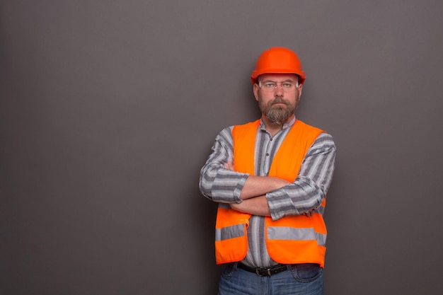 Free photo profile of hipster bearded builder with his arms crossed. man in orange helmet and protective jacket posing seriously on dark grey.