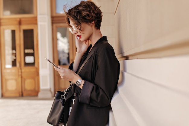 Free Photo profile photo of cool woman with short hair in black jacket holds tablet outdoors. beautifuul woman in eyeglases with handbag posing at street.