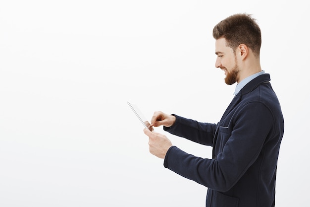 Profile shot of determined confident and successful stylish businessman with beard and awesome hairstyle in elegant suit using digital tablet smiling delighted checking income of company