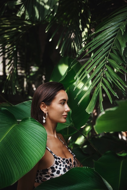 Free photo profile of tanned woman in a bikini, wearing earrings, standing in jungle tropical leaves and palm trees.