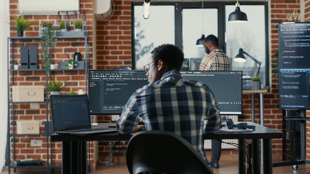 Free photo programer coding on laptop sitting down at desk with computer screens parsing code in software agency. software developer compiling algorithms with cloud programers working in the background.