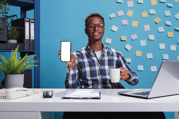 Project manager pointing phone device with mockup display to camera. Young adult business person sitting in company modern workspace office while showing white screen isolated template to camera.