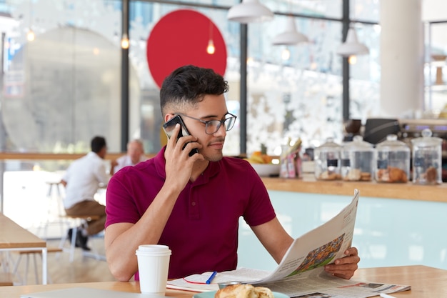 Prosperous attractive businessman writes latest issue of newspaper, focused on interesting publication during coffee break, discusses something with partner via cell phone, writes in notebook