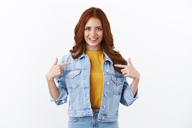 Proud and confident, motivated cute ginger girl with freckles in denim jacket, pointing herself and smiling boastful, feeling honored and self-assured, suggest help, accomplish goal, white wall