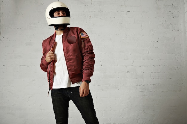 Free photo proud looking motorbiker in a plain white helmet, bordeaux nylon bomber jacket, jeans and t-shirt against white wall background