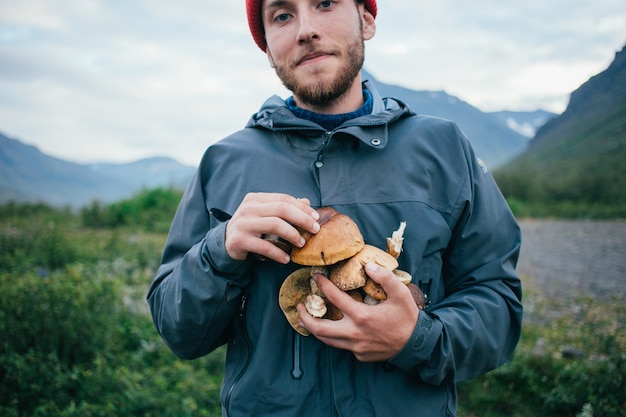 Free Photo proud picker man in traditional blue wool sweater with ornaments stands on camping ground in mountains, holds in arms pile of delicious and organic mushrooms