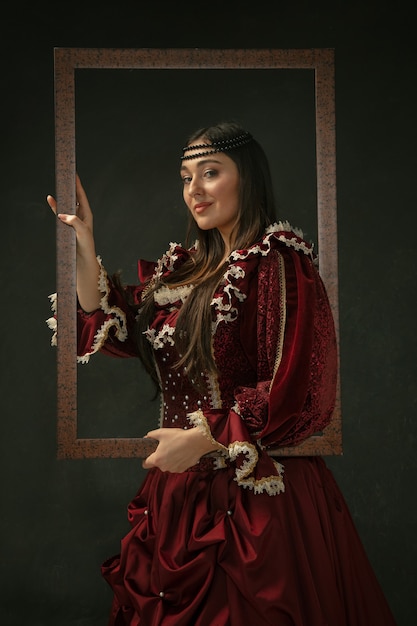 Free photo proud. portrait of medieval young woman in red vintage clothing standing on dark background. female model as a duchess, royal person. concept of comparison of eras, modern, fashion, beauty.