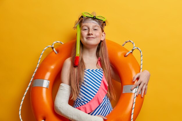 Free Photo proud satisfied redhead girl poses in inflated lifebuoy, wears snorkeling mask and swimsuit, enjoys swimming in sea, has broken arm in cast after getting into accident, stands against yellow wall