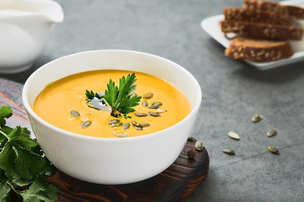 Pumpkin and carrot chowder with olive oil in a white cup, pumpkin seeds and parsley on a gray background. Close-up on seasonal vegetarian soup, selective sharpening on parsley