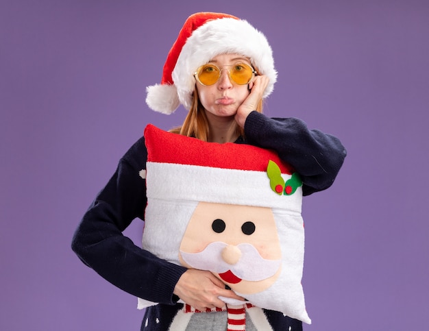 Free photo pursing lips young beautiful girl wearing christmas sweater and hat with glasses holding christmas pillow putting hand on cheek isolated on purple wall