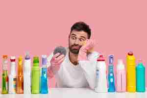 Free photo puzzled bearded man looks with stupefied expression at sponge, wears protective gloves, sits at desk with cleaning agents