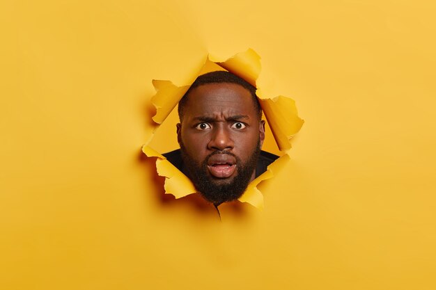 Puzzled black man with thiсk bristle, looks with angry surprised face expression, keeps head in torn paper hole, stands bothered and disappointed. Yellow background