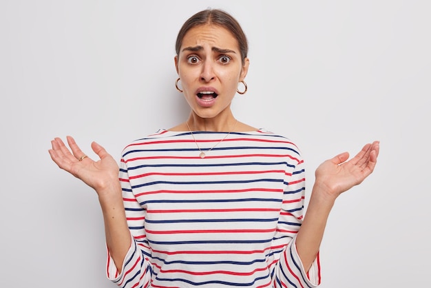 Free Photo puzzled brunette woman spreads palms and looks indignant looks displeased wears casual striped jumper isolated over white background. negaive human reactions. embarrassed female model indoor