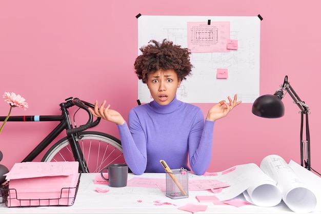 Free Photo puzzled clueless young woman spreads palms creats architectural project make research poses at desktop dressed casually