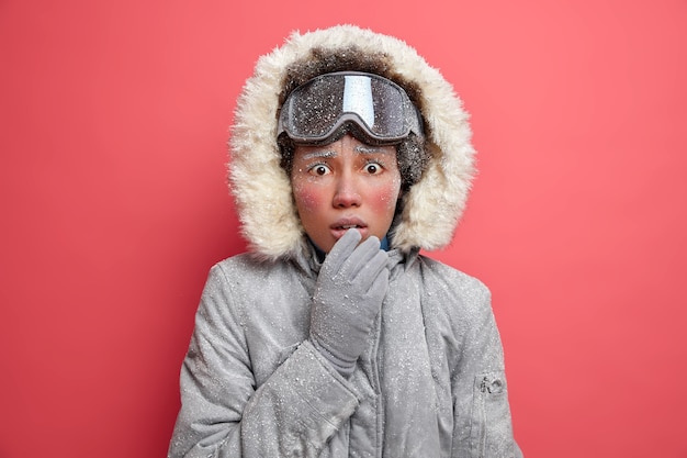 Free Photo puzzled frozen woman with red icy face looks embarrassed trembles from cold wears ski goggles grey coat goes hiking in stormy snow weather