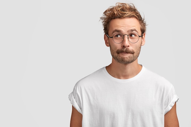 Puzzled handsome man with mustache, bites lower lip and looks curiously aside, thinks about something, dressed in casual white t shirt, stands against blank wall