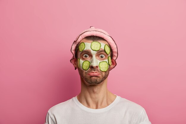 Free photo puzzled male model applies clay facial mask and slices of cucumber ro rejuvenate skin, wears bath hat, white t shirt