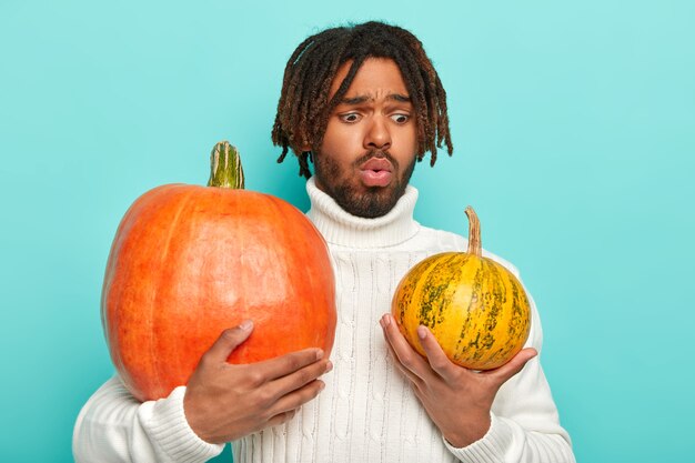 Puzzled man holds two pumpkins, chooses which better for preparing healthy nutritious meal, wears white sweater with long collar