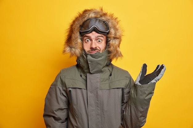 Puzzled man in winter outfit raises hand and looks confused wears hood of his anorak coat goes skiing during spare time.