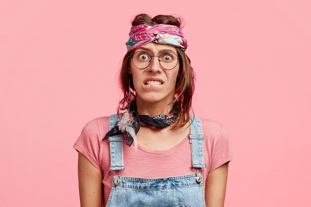 Puzzled nervous hippy woman bites lower lips, looks in embarrassement, worries before important event, wears stylish bandana and dungarees