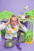 Free photo puzzled overwhelmed elderly man in spectacles has much to do wears domestic clothes with stuck reminding sticky notes keeps hands on head sits on floor near office desk piles of cut green paper around