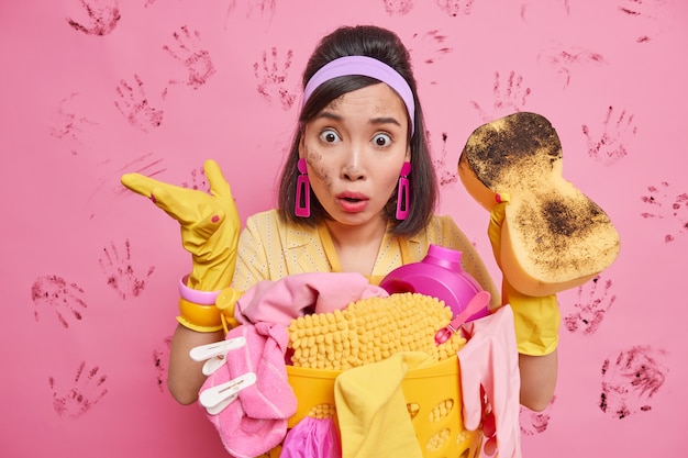 Free photo puzzled shocked brunette young asian woman wears headband rubber protective gloves stunned to see such mess and chaos in room much dirt holds sponge to reduce dirt stands near laundry basket