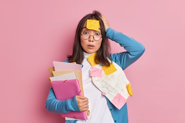 Puzzled shocked female office worker overloaded with paper work stunned to have deadline for finishing research holds folders keeps hand on head wears round spectacles.