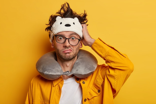 Free Photo puzzled tourist scratches head and considers his mistake, looks thorugh glasses, wears neckpillow for traveling in transport