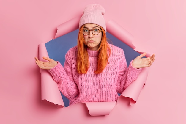 Free photo puzzled unaware redhead woman spreads palms with clueless expression purses lips and being unaware cannot decide what to do wears fashionable pink hat and knitted sweater breaks through paper