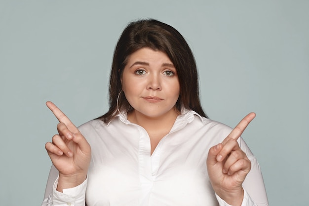 Free photo puzzled young dark haired businesswoman with chubby cheeks and curvy body pointing her index fingers at opposite directions, can't choose between two business concepts, having doubtful look