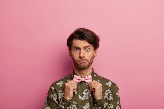 Free Photo puzzled young guy raises eyebrows, has bristle, adjustes bowtie, wears fashionable shirt with modern print, dresses tastefully, stands against rosy space. people