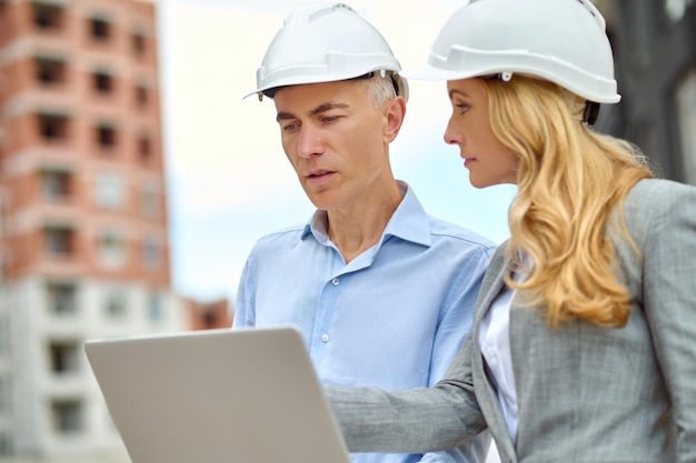 Free photo question. profile of demanding business woman with long blond hair in protective helmet looking attentively at talking man with laptop standing at construction site